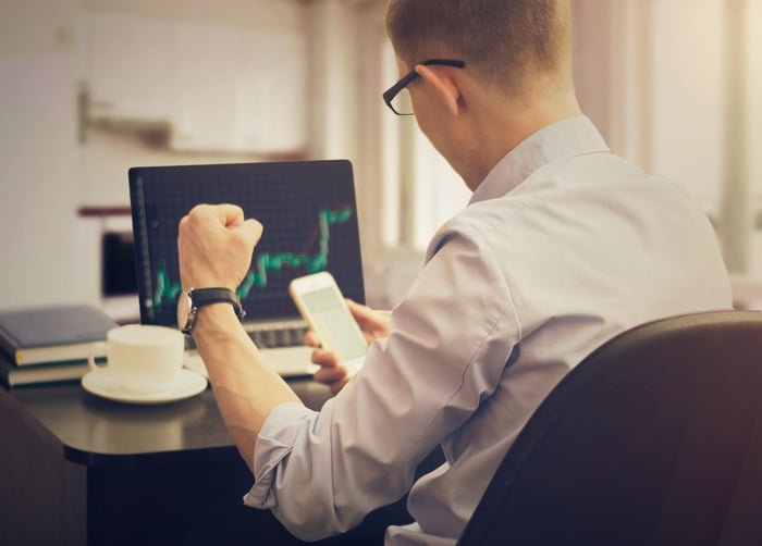 An investor seen from behind at a desk cheers while looking at a phone.